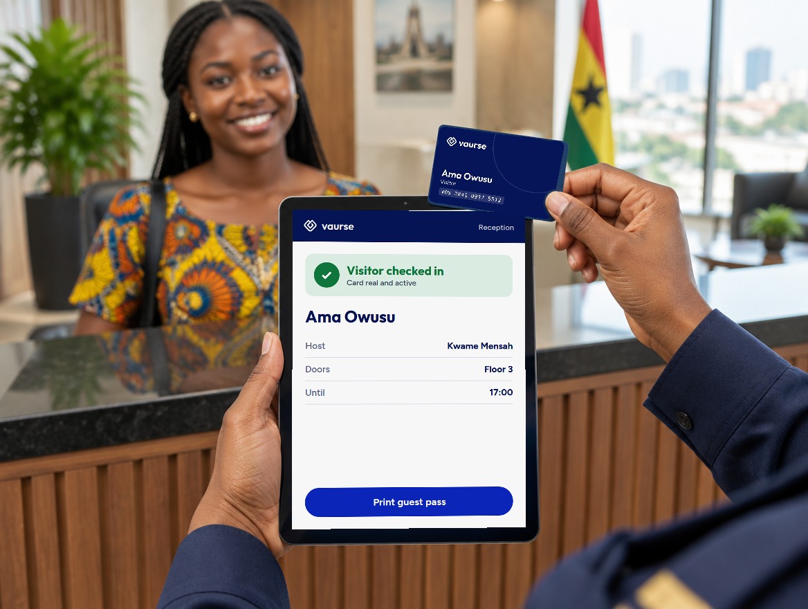 At an office reception, a guard holds up a tablet showing Visitor checked in, Ama Owusu, and the visitor's navy Voscard, while Ama smiles at the desk behind.